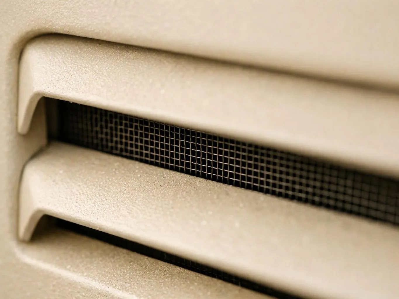 Close-up of a beige metal vent with horizontal slats and a dark mesh screen behind them